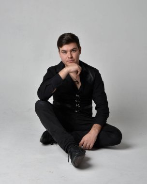 Siyah gömlek ve gotik yelek giyen esmer bir adamın tam boy portresi. Gri bir stüdyonun arka planında izole bir poz..