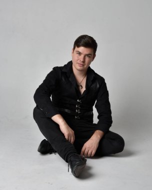 Siyah gömlek ve gotik yelek giyen esmer bir adamın tam boy portresi. Gri bir stüdyonun arka planında izole bir poz..