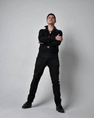 Siyah gömlek ve gotik yelek giyen esmer bir adamın tam boy portresi. Gri bir stüdyonun arka planında izole bir şekilde duruyor..