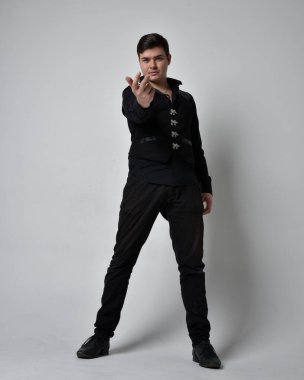 Siyah gömlek ve gotik yelek giyen esmer bir adamın tam boy portresi. Gri bir stüdyonun arka planında izole bir şekilde duruyor..