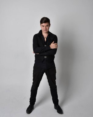 Siyah gömlek ve gotik yelek giyen esmer bir adamın tam boy portresi. Gri bir stüdyonun arka planında izole bir şekilde duruyor..