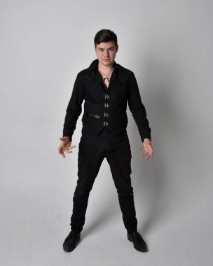 Siyah gömlek ve gotik yelek giyen esmer bir adamın tam boy portresi. Gri bir stüdyonun arka planında izole bir şekilde duruyor..