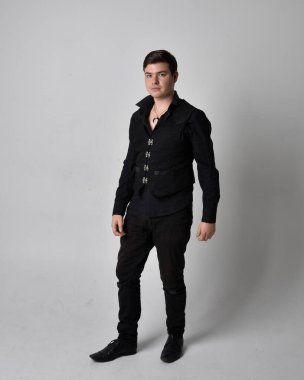 Siyah gömlek ve gotik yelek giyen esmer bir adamın tam boy portresi. Gri bir stüdyonun arka planında izole bir şekilde duruyor..