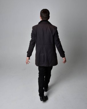 Siyah deri ceket giyen esmer bir adamın tam boy portresi. Geriye dönük poz vermek, kameradan uzaklaşmak gri bir stüdyonun arka planında izole edilmiş..