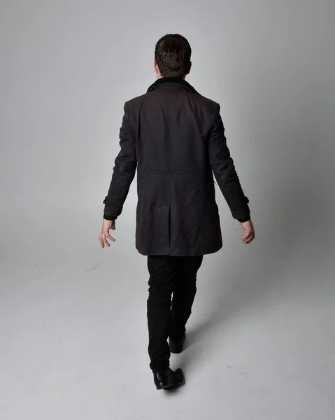 Full length portrait of a brunette man wearing black leather coat.  Standing pose facing backwards, walking away from the camera isolated  against a grey studio background.