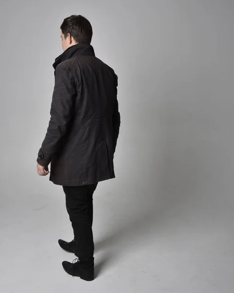 黒革のコートを着たブルネットの男の完全な長さの肖像画 後ろ向きに立ち 灰色のスタジオの背景に隔離されたカメラから離れて歩くポーズ — ストック写真