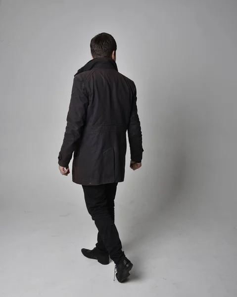 黒革のコートを着たブルネットの男の完全な長さの肖像画 後ろ向きに立ち 灰色のスタジオの背景に隔離されたカメラから離れて歩くポーズ — ストック写真