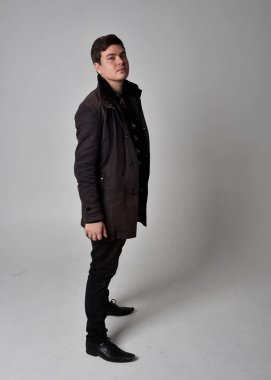 Siyah deri ceket giyen esmer bir adamın tam boy portresi. Gri bir stüdyonun arka planında izole bir şekilde duruyor..