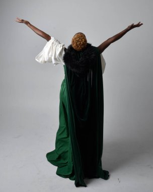 Uzun yeşil ortaçağ fantezi elbisesi ve kadife pelerin giyen, açık gri bir stüdyo arka planında oturan Afrikalı güzel bir kadının tam boy portresi..