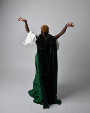 Uzun yeşil ortaçağ fantezi elbisesi ve kadife pelerin giyen, açık gri bir stüdyo arka planında oturan Afrikalı güzel bir kadının tam boy portresi..