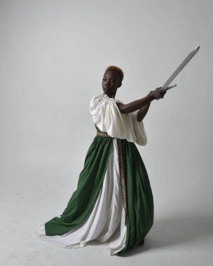 Uzun yeşil bir ortaçağ fantezi elbisesi giymiş, açık gri bir stüdyo arka planında ayakta duran güzel Afrikalı kadının uzun bir portresi..