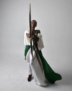 Uzun yeşil bir ortaçağ fantezi elbisesi giymiş, elinde ok ve yay tutan, açık gri bir stüdyo arka planında ayakta duran güzel Afrikalı kadının tam boy portresi..