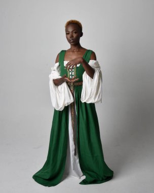 Uzun yeşil bir ortaçağ fantezi elbisesi giyen, açık gri bir stüdyo arka planında ayakta duran ve dans eden güzel Afrikalı kadının tam boy portresi..