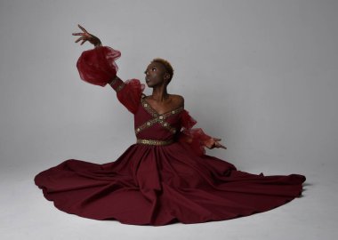 Uzun kırmızı rönesans fantezi elbisesi giyen, açık gri bir stüdyo arka planında oturan Afrikalı güzel bir kadının tam boy portresi..