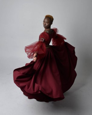 Uzun kırmızı rönesans fantezi elbisesi giyen, açık gri stüdyo arka planında duran Afrikalı güzel bir kadının tam boy portresi..