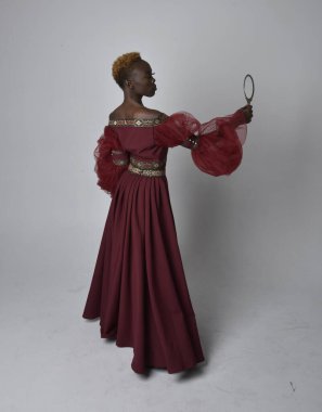 Uzun kırmızı rönesans fantezi elbisesi giyen, açık gri stüdyo arka planında duran Afrikalı güzel bir kadının tam boy portresi..