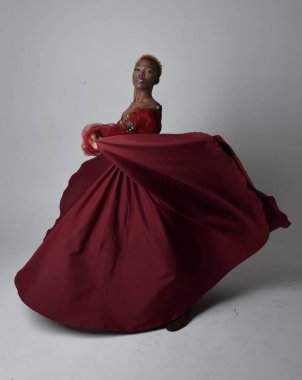 Uzun kırmızı rönesans fantezi elbisesi giyen, açık gri stüdyo arka planında duran Afrikalı güzel bir kadının tam boy portresi..