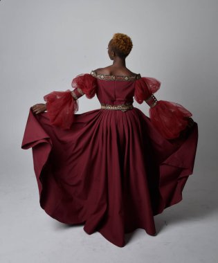 Uzun kırmızı rönesans fantezi elbisesi giyen, açık gri stüdyo arka planında duran Afrikalı güzel bir kadının tam boy portresi..