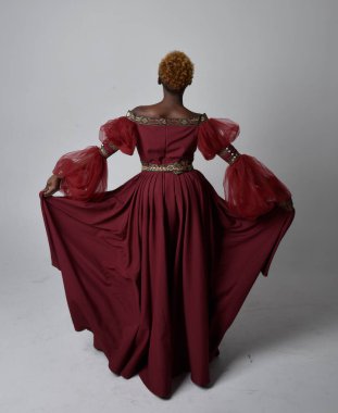 Uzun kırmızı rönesans fantezi elbisesi giyen, açık gri stüdyo arka planında duran Afrikalı güzel bir kadının tam boy portresi..