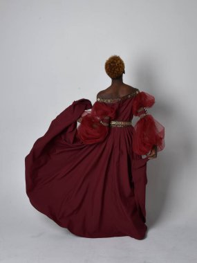 Uzun kırmızı rönesans fantezi elbisesi giyen, açık gri stüdyo arka planında duran Afrikalı güzel bir kadının tam boy portresi..