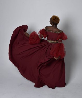 Uzun kırmızı rönesans fantezi elbisesi giyen, açık gri stüdyo arka planında duran Afrikalı güzel bir kadının tam boy portresi..