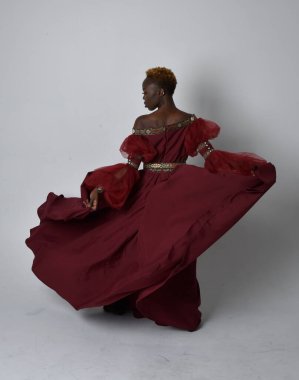 Uzun kırmızı rönesans fantezi elbisesi giyen, açık gri stüdyo arka planında duran Afrikalı güzel bir kadının tam boy portresi..