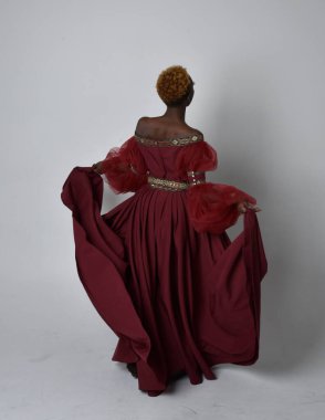 Uzun kırmızı rönesans fantezi elbisesi giyen, açık gri stüdyo arka planında duran Afrikalı güzel bir kadının tam boy portresi..