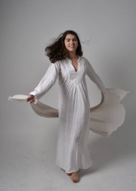 Klasik beyaz önlük giyen ve başörtüsünü İncil tarzında örten, hafif stüdyo arka planında duran genç bir kadının tam boy portresi..