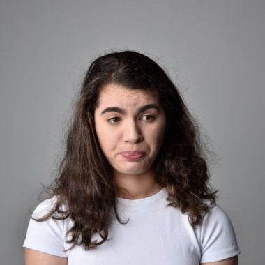 Açık gri stüdyo arka planındaki yüz ifadelerinin üzerinde, doğal kahverengi saçlı genç bir kadının yakın portresi var..