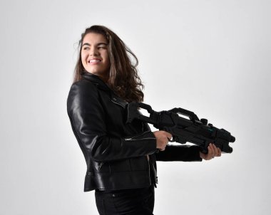 Doğal kahverengi saçlı, korseli, siyah deri bilimkurgu kıyafetli açık gri stüdyo arka planında duran genç bir kadının tam boy portresi..