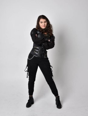 Doğal kahverengi saçlı, korseli, siyah deri bilimkurgu kıyafetli açık gri stüdyo arka planında duran genç bir kadının tam boy portresi..