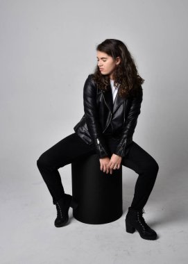 Doğal kahverengi saçlı, ceketli, siyah deri elbiseli, açık renk stüdyo arka planında oturan genç bir kadının tam boy portresi..