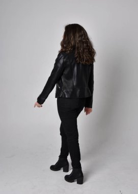 Doğal kahverengi saçlı, ceketli, siyah deri bilimkurgu kıyafetli, açık renk stüdyo arka planında kameraya dönük duran genç bir kadının tam boy portresi..