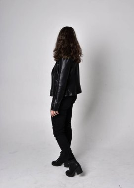 Doğal kahverengi saçlı, ceketli, siyah deri bilimkurgu kıyafetli, açık renk stüdyo arka planında kameraya dönük duran genç bir kadının tam boy portresi..