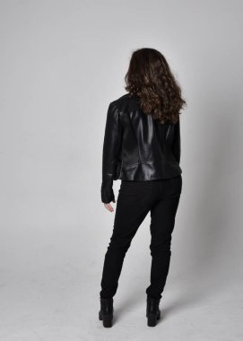 Doğal kahverengi saçlı, ceketli, siyah deri bilimkurgu kıyafetli, açık renk stüdyo arka planında kameraya dönük duran genç bir kadının tam boy portresi..