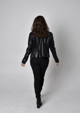 Doğal kahverengi saçlı, ceketli, siyah deri bilimkurgu kıyafetli, açık renk stüdyo arka planında kameraya dönük duran genç bir kadının tam boy portresi..