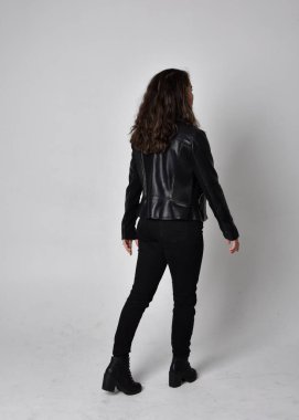 Doğal kahverengi saçlı, ceketli, siyah deri bilimkurgu kıyafetli, açık renk stüdyo arka planında kameraya dönük duran genç bir kadının tam boy portresi..