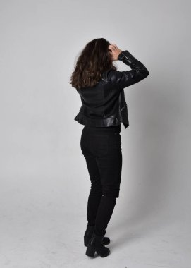 Doğal kahverengi saçlı, ceketli, siyah deri bilimkurgu kıyafetli, açık renk stüdyo arka planında kameraya dönük duran genç bir kadının tam boy portresi..