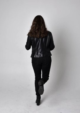 Doğal kahverengi saçlı, ceketli, siyah deri bilimkurgu kıyafetli, açık renk stüdyo arka planında kameraya dönük duran genç bir kadının tam boy portresi..