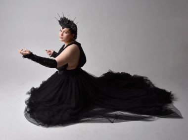 Uzun boylu, kısa saçlı, uzun siyah smokinli, taç ve eldivenli, açık renk stüdyo arka planında poz veren genç ve iri yarı bir kadının portresi..