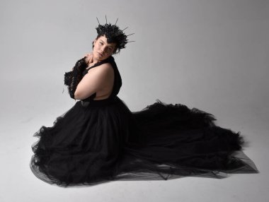 Uzun boylu, kısa saçlı, uzun siyah smokinli, taç ve eldivenli, açık renk stüdyo arka planında poz veren genç ve iri yarı bir kadının portresi..