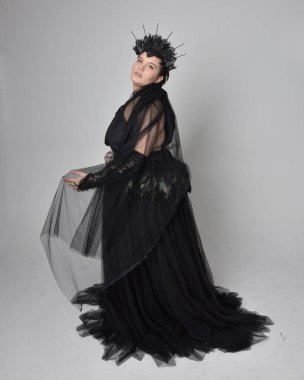 Uzun boylu, kısa saçlı, uzun siyah tül gotik cüppeli, taç ve eldivenli, açık renk stüdyo arka planında duran genç bir kadının portresi..