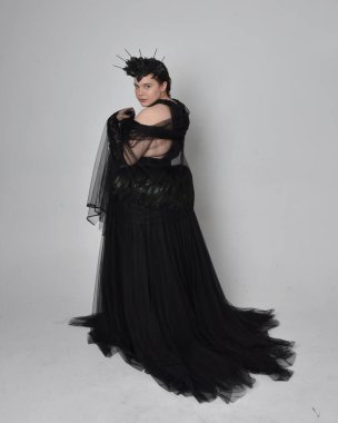 Uzun boylu, kısa saçlı, uzun siyah tül gotik cüppeli, taç ve eldivenli, açık renk stüdyo arka planında duran genç bir kadının portresi..