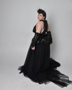 Uzun boylu, kısa saçlı, uzun siyah tül gotik cüppeli, taç ve eldivenli, açık renk stüdyo arka planında duran genç bir kadının portresi..