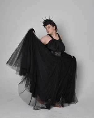 Uzun boylu, kısa saçlı, uzun siyah tül gotik cüppeli, taç ve eldivenli, açık renk stüdyo arka planında duran genç bir kadının portresi..