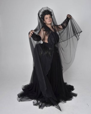 Uzun boylu, kısa saçlı, uzun siyah tül gotik cüppeli, karga, eldiven ve peçeli, açık renk stüdyo arka planında duran, uzun boylu bir kadının portresi..