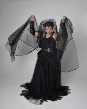 Uzun boylu, kısa saçlı, uzun siyah tül gotik cüppeli, karga, eldiven ve peçeli, açık renk stüdyo arka planında duran, uzun boylu bir kadının portresi..