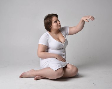 Uzun boylu, esmer, kısa saçlı, dar beyaz beden elbisesi giyen, yerde hareketli pozlar veren, hafif stüdyo arka planlı genç bir kadının portresi..