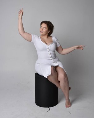 Full length portrait of young plus sized woman with short brunette hair,  wearing tight white body con dress,   sitting pose on chair with gestural hands with light studio background. clipart