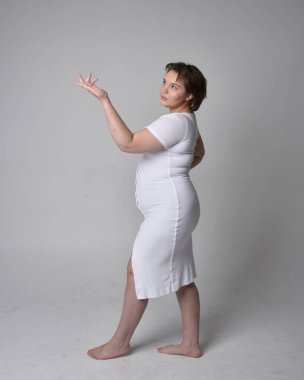 Uzun boylu, esmer kısa saçlı, dar beyaz bir vücut elbisesi giyen, hafif stüdyo arka planda ayakta duran genç bir kadının portresi..